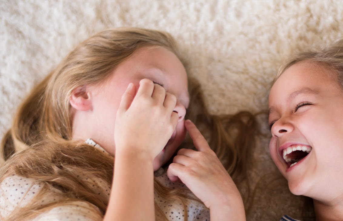 Två flickor ligger på en ljus matta och skrattar.