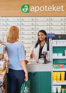 En kvinnlig kund står vid disken på ett apotek och pratar med en apotekare. Kunden ser ut att ställa frågor eller få rådgivning om mediciner.