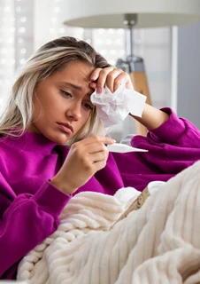 En kvinna med feber sitter i en soffa och har precis använt en febertermometer.