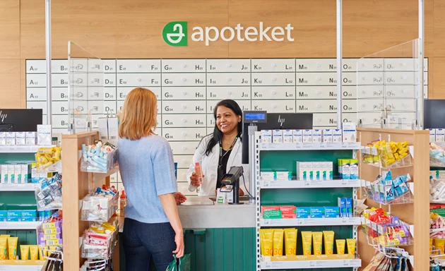 En kvinnlig kund står vid disken på ett apotek och pratar med en apotekare. Kunden ser ut att ställa frågor eller få rådgivning om mediciner.