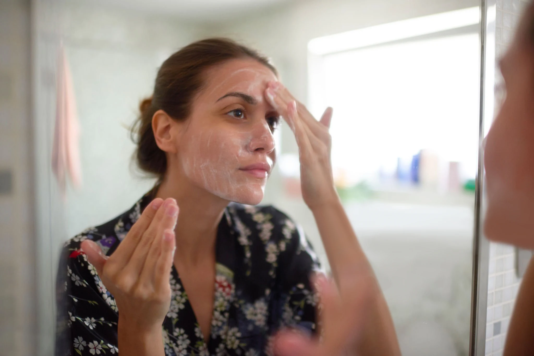 En kvinna står framför badrumsspegeln och applicera en rengöring eller ansiktsmask på sitt ansikte.