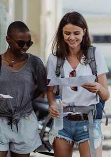 Två kvinnor går längs en gata och kollar ner på kartor de håller i handen under en sommardag.