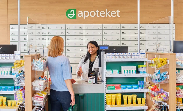 En kvinnlig kund står vid disken på ett apotek och pratar med en apotekare. Kunden ser ut att ställa frågor eller få rådgivning om mediciner.