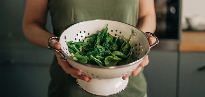 Kvinna i grönt linne håller ett durkslag med nysköljd färsk spenat.
