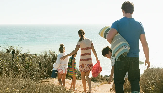En familj går på en stig ner till en strand. I händerna håller de handdukar och badväskor.