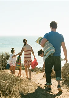 En familj går på en stig ner till en strand. I händerna håller de handdukar och badväskor.