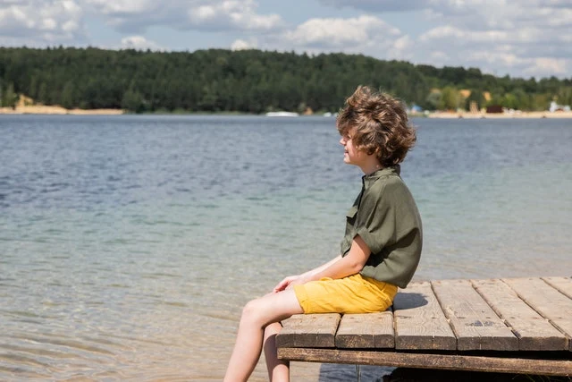En pojke sitter på en brygga och tittar ut över en sjö under en sommardag. 