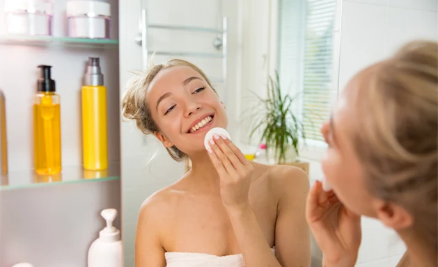 En kvinna står framför spegeln i badrummet med en bomullspad i handen, tittandes på sin reflektion medan hon rengör sin haka