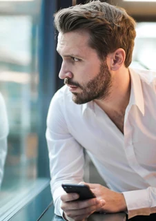 En man tittar ut genom ett fönster och håller en mobiltelefon i ena handen. 