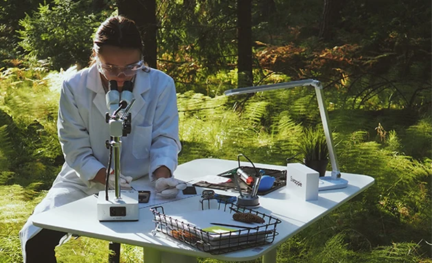 En farmaceut sitter ute i skogen och tittar genom ett mikroskåp.