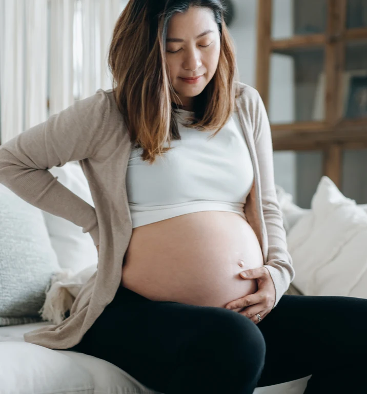 En gravid kvinna sitter på en soffa med ena handen på magen och den andra på ryggen. 