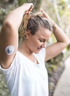 Kvinna står utomhus och sätter upp sitt hår med en realtids-CGM på underarmen.
