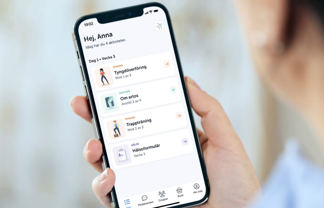 En hand som håller i en mobiltelefon. På telefonens skärm syns Joint Academys app med övningar.
