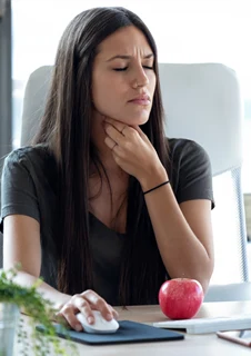 En kvinna sitter vid ett skrivbord och håller ena handen framför halsen. 