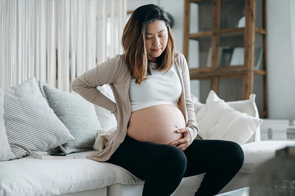 En gravid kvinna sitter i en soffa och håller ena handen på magen och den andra på ryggen. 