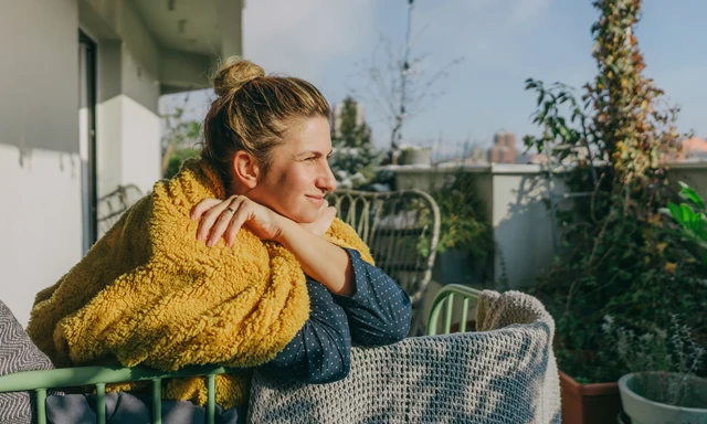 En kvinna inlindad i en filt sitter på en balkong och njuter av solen. 