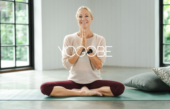 En kvinna sitter på en yogamatta i en hälsningsposition och ska träna yoga tillsammans med Yogobe.