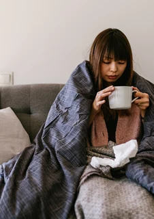 En kvinna med influensa sitter i en soffa inlindad i filtar och dricker en varm dryck.