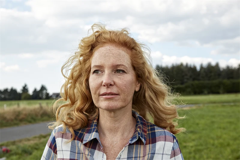 En medelålders kvinna med rött hår står utomhus med en landsväg och skog bakom sig 