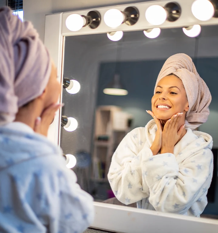 En kvinna med morgonrock och en handduk på huvudet tittar sig själv i spegeln och ler. 