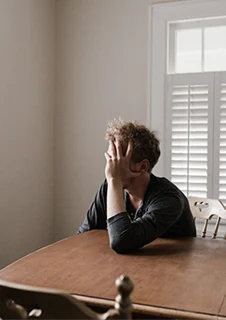 En man sitter vid ett bord med handen framför ansiktet och tittar ut genom ett fönster. 