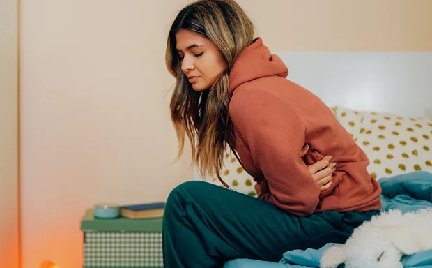 En kvinna med mensvärk sitter på sängkanten och håller om magen med båda händerna. 