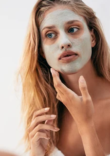 En kvinna i ett badrum smörjer försiktigt på en ansiktsmask med en subtil grön ton. 