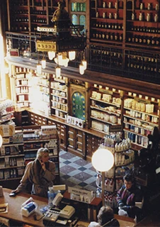 Ett gammaldags apotek med trähyllor fulla av produkter, där en gyllene lejonstaty pryder rummet ovanifrån. Kunder står vid disken och handlar.