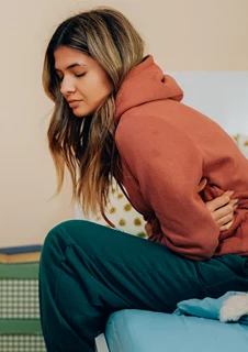 En kvinna med mensvärk sitter på sängkanten och håller om magen med båda händerna. 