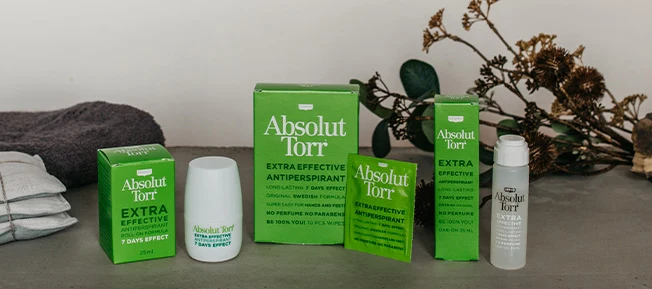 Både Produkter och ytterförpackningar av Roll-on, Dab-on och servetter från Absolut Torr placerade bredvid varandra mot en grå bänkskiva med en torkad växt och handduk i bakgrunden.