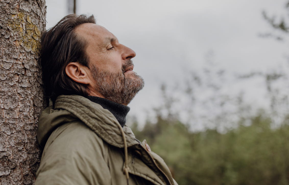 En medelålders man i grön jacka lutar sig mot en tall ute i skogen.