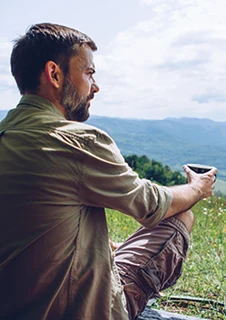 En man sitter i ett tält med öppen öppning. Man ser hans ryggtavla och utsikt över skog och berg.