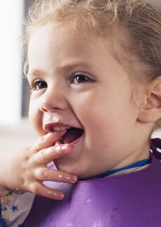Ett barn med en haklapp sitter med matrester på händerna och stoppar dem i munnen.