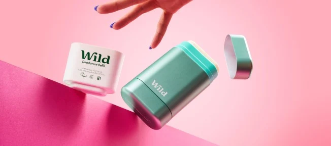 Wild deodorant och refill i luften mot rosa bakgrund, med en hand som sträcker sig mot produkterna.