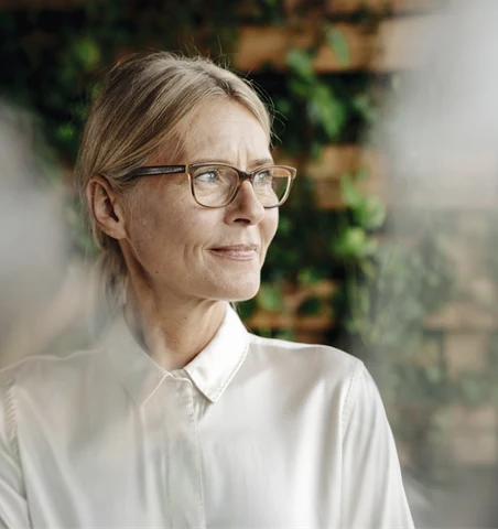 En medelålders kvinna med glasögon och vit skjorta tittar ut genom ett fönster. 