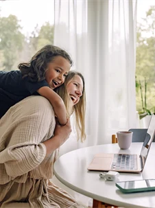 Glad pojke kramar om sin mamma som sitter på en stol vid ett bord hemma.