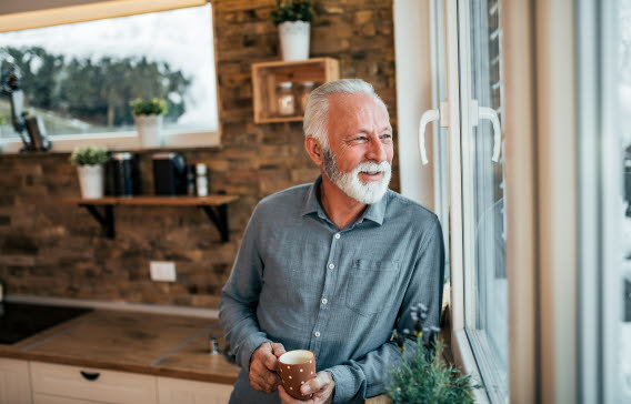En äldre man tittar ut genom ett fönster, i händerna håller han en kaffekopp.