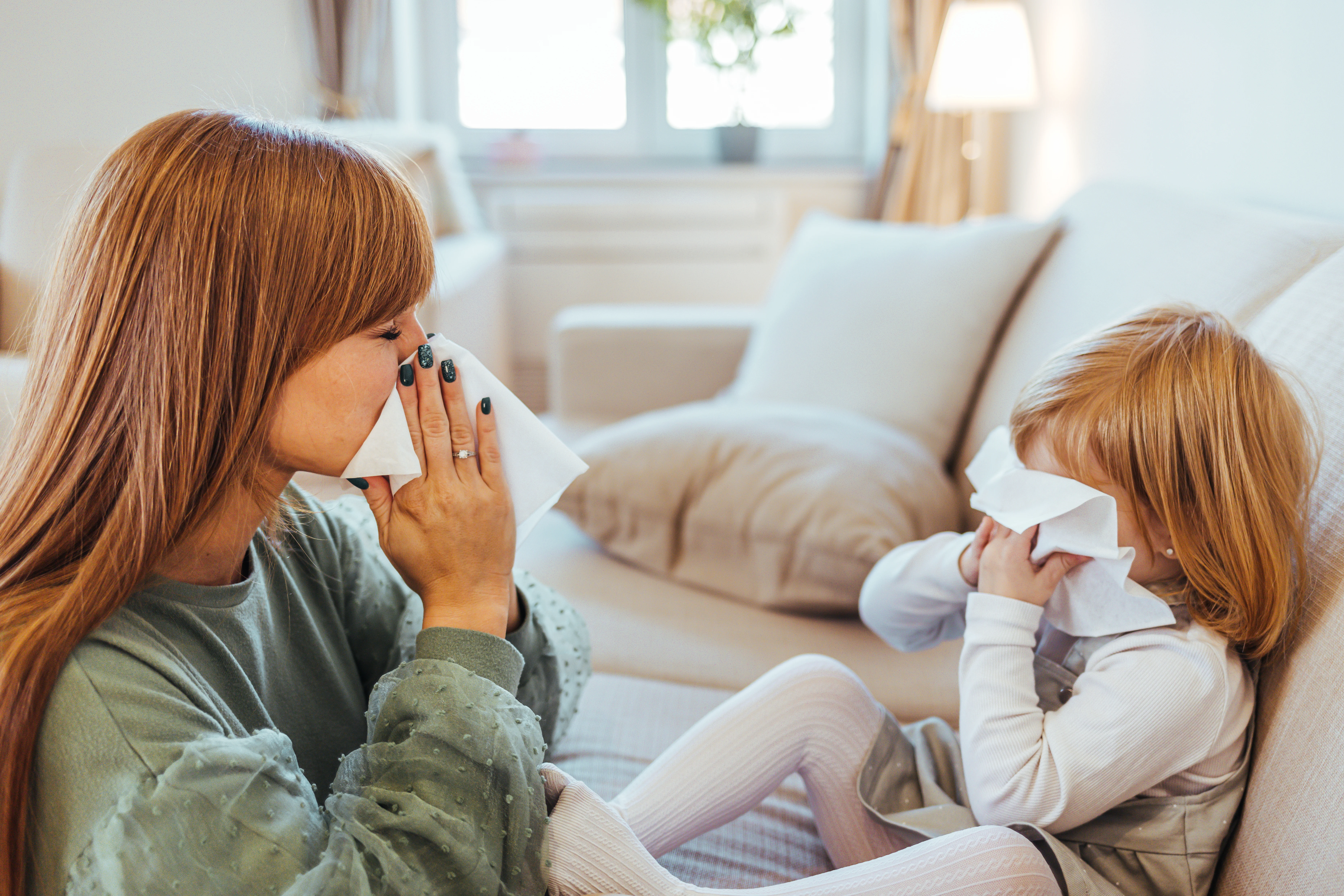 En kvinna och ett litet barn sitter på en soffa och snyter sig samtidigt med vita pappersservetter. 