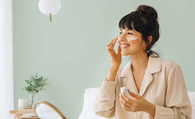 En kvinna sitter i sin säng omgiven av hudvårdsprodukter i ett harmoniskt rum. Hon applicerar hudkräm på sina kinder medan hon blickar ut genom fönstret