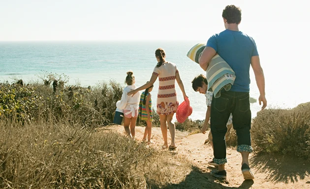 En familj går på en stig ner till en strand. I händerna håller de handdukar och badväskor.