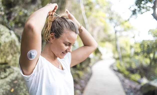 Kvinna står utomhus och sätter upp sitt hår med en realtids-CGM på underarmen.