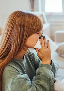 En kvinna och ett litet barn sitter på en soffa och snyter sig samtidigt med vita pappersservetter. 